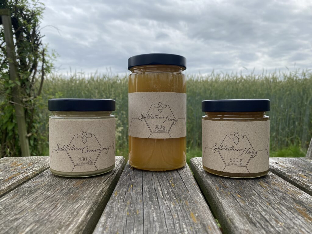 Drei volle Honiggläser, die auf einer Holzplattform stehen. Im Hintergrund ein grünes Feld und ein bewölkter Himmel.